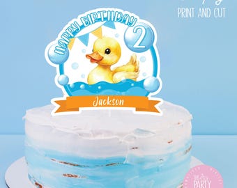 May include: A birthday cake with a blue and white ombre design, topped with a duck-themed cake topper. The topper features a yellow duck, the words "Happy Birthday 2", and the name "Jackson". The words "Simply Print and Cut" are also visible.