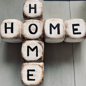 May include: Wooden blocks spelling out the word "HOME" in black paint on a white background. The blocks are arranged in a cross shape.