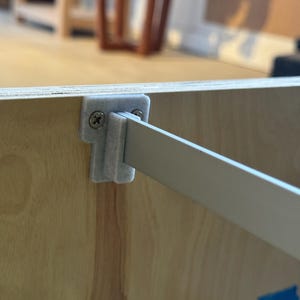 Puede incluir: Primer plano de un panel de madera de color claro con una viga de soporte blanca fijada con un soporte de plástico gris y tornillos. La veta de la madera es visible. Un trozo de cinta azul con escritura es visible.