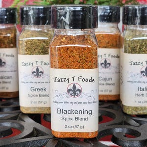May include: Several spice jars with black lids and labels reading "Jazzy T Foods". The jars contain different spice blends, including "Blackening Spice Blend" (57g), "Greek Spice Blend" (57g), and "Italian Herb Blend" (28.35g).