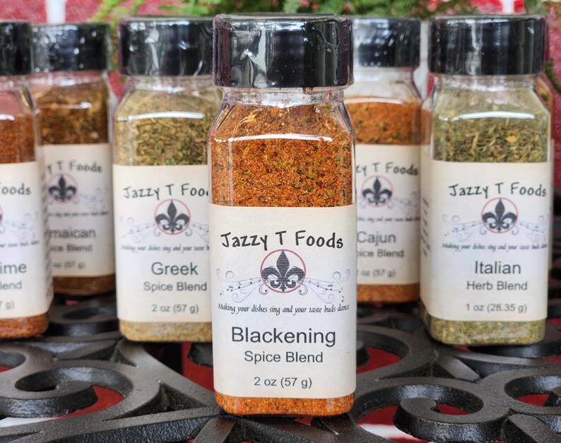 May include: Several spice jars with black lids and labels reading "Jazzy T Foods". The jars contain different spice blends, including "Blackening Spice Blend" (57g), "Greek Spice Blend" (57g), and "Italian Herb Blend" (28.35g).
