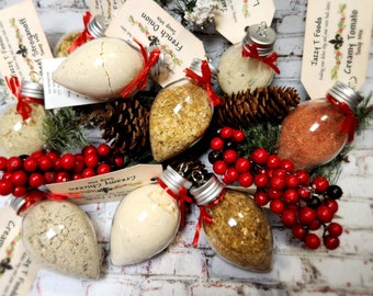 May include: A collection of clear, bulb-shaped ornaments filled with various dry soup mixes, each topped with a silver cap, a red ribbon, and a beige tag labeled Jazzy T Foods with specific soup names like French Onion and Creamy Tomato.