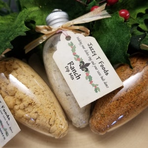 May include: Three glass bottles filled with dip mix, each with a silver cap and a decorative tag. The tags read "Jazzy T Foods" and list the dip mix flavors: Ranch, Bacon Cheese Habanero, and another unreadable flavor. The bottles are tied with raffia.