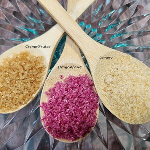 May include: Three wooden spoons filled with different colored sugar crystals. The spoons are labeled "Creme Brulee", "Dragonfruit", and "Lemon".