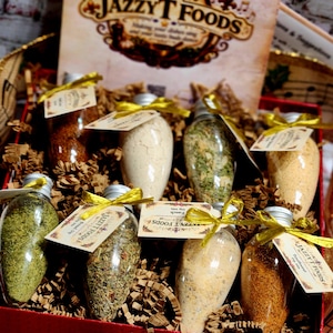 May include: A gift box containing eight clear, bulb-shaped glass jars filled with various dry spice and dip mixes. Each jar has a silver screw-top lid, a gold ribbon, and a tag labeled Jazzy T Foods. The box includes a branded display card.