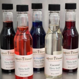 May include: Five clear glass bottles with black caps, each filled with a different colored flavored syrup. Flavors include Cherry, Peach, Blue Raspberry, Coconut, and Strawberry. Each bottle has a label that reads "Jazzy Foods Dirty Soda Flavored Syrup" and lists the flavor and size, 5 fl oz (148ml).