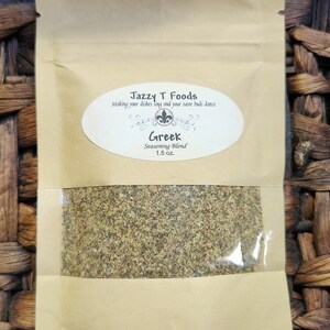 May include: A brown paper pouch with a clear plastic window showing a green Greek seasoning blend. The pouch has a white label with the text "Jazzy T Foods" and "Making your dishes sing and your taste buds dance". The label also has a white oval with the text "Greek Seasoning Blend 1.5 oz."
