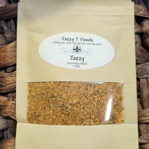 May include: A brown paper pouch with a clear plastic window showing a yellow-brown seasoning blend. The pouch has a white label with the text "Jazzy T Foods" and "Making your dishes sing and your taste buds dance". Below this is a smaller white label with the text "Jazzy Seasoning Blend 1.5oz."