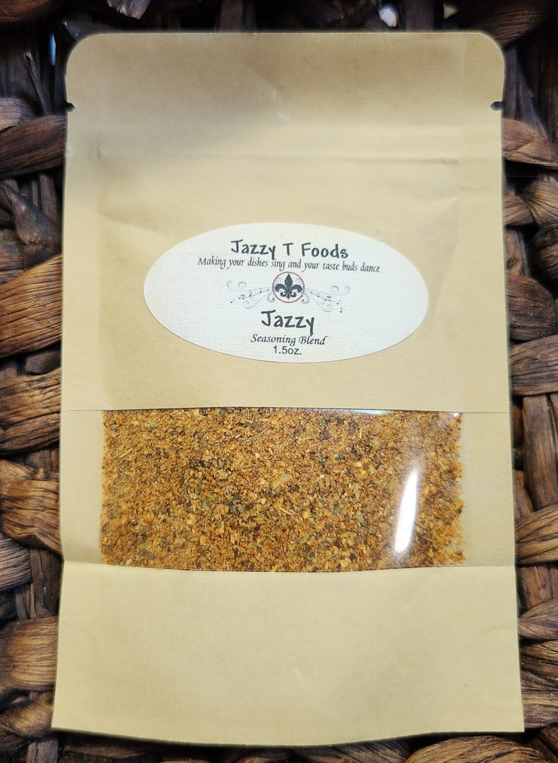 May include: A brown paper pouch with a clear plastic window showing a yellow-brown seasoning blend. The pouch has a white label with the text "Jazzy T Foods" and "Making your dishes sing and your taste buds dance". Below this is a smaller white label with the text "Jazzy Seasoning Blend 1.5oz."