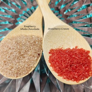 May include: Two wooden spoons filled with red and brown sugar crystals. The spoon on the left is labeled "Raspberry White Chocolate" and the spoon on the right is labeled "Strawberry Cream".