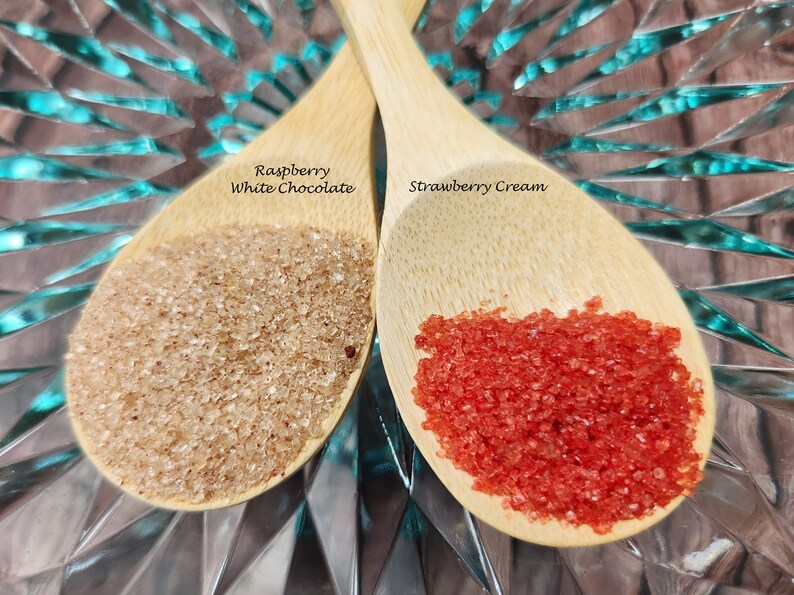 May include: Two wooden spoons filled with red and brown sugar crystals. The spoon on the left is labeled "Raspberry White Chocolate" and the spoon on the right is labeled "Strawberry Cream".