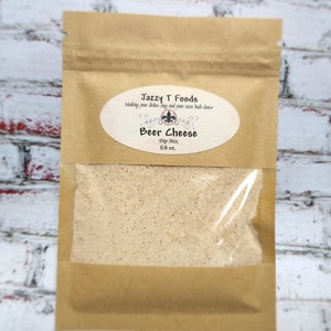 May include: A brown paper bag with a clear window, containing a beige dip mix. The bag has an oval label that reads "Jazzy T Foods Beer Cheese Dip Mix 0.6 oz."