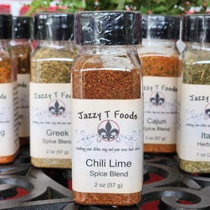 May include: A selection of spice blends in clear glass jars with black lids. The labels read "Jazzy T Foods" and include "Chili Lime", "Greek", "Cajun", "Italian Herb Blend", and "Blackening" spice blends. Each jar contains 57 g of spice.