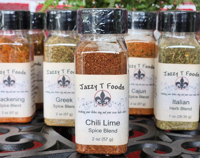 May include: A selection of spice blends in clear glass jars with black lids. The labels read "Jazzy T Foods" and include "Chili Lime", "Greek", "Cajun", "Italian Herb Blend", and "Blackening" spice blends. Each jar contains 57 g of spice.