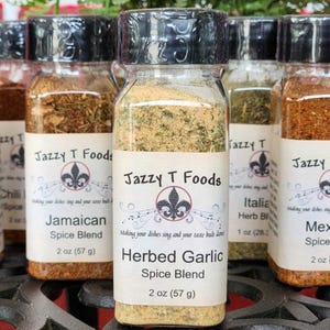 May include: A selection of glass spice jars with black lids, each labelled with a different spice blend name. The labels feature the text "Jazzy T Foods" and a fleur-de-lis design. The jars contain various dried herbs and spices, including a herbed garlic blend.