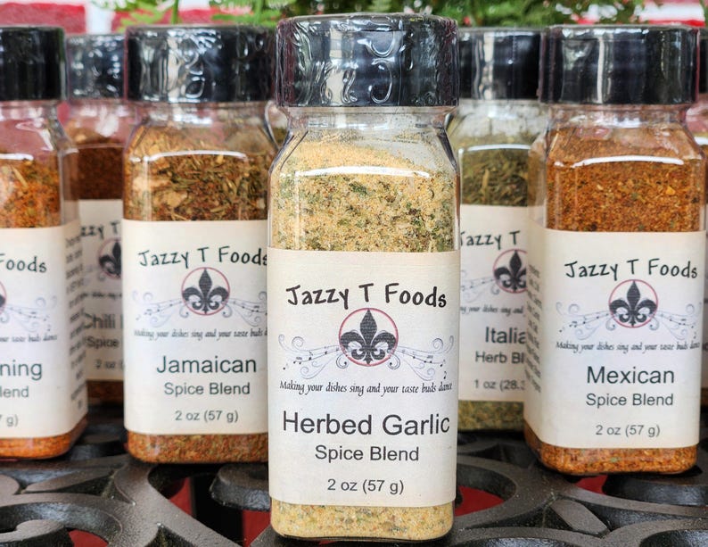 May include: A selection of glass spice jars with black lids, each labelled with a different spice blend name. The labels feature the text "Jazzy T Foods" and a fleur-de-lis design. The jars contain various dried herbs and spices, including a herbed garlic blend.