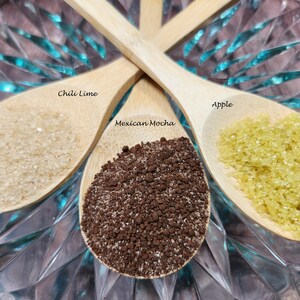 May include: Three wooden spoons filled with different ingredients. The spoon on the left is filled with white sugar and labeled "Chili Lime". The spoon in the middle is filled with brown coffee grounds and labeled "Mexican Mocha". The spoon on the right is filled with yellow sugar and labeled "Apple".