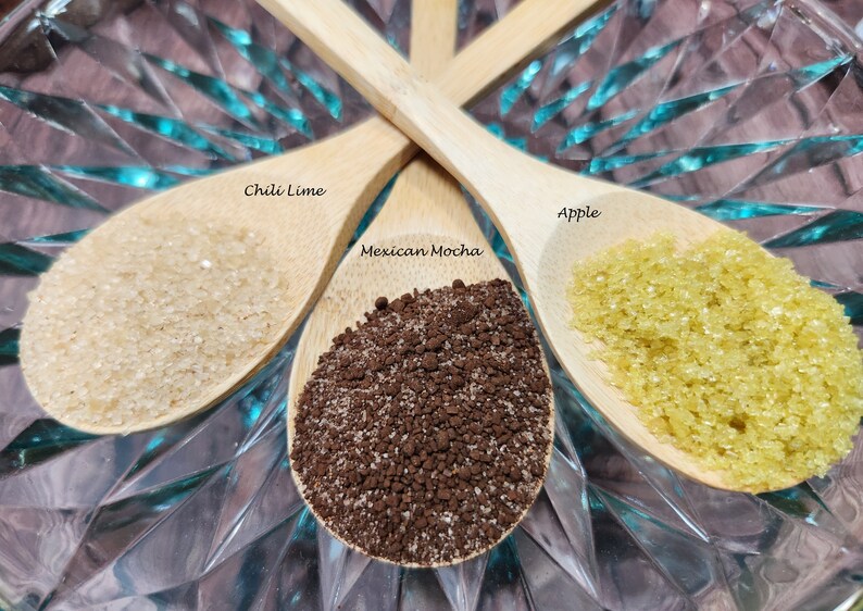 May include: Three wooden spoons filled with different ingredients. The spoon on the left is filled with white sugar and labeled "Chili Lime". The spoon in the middle is filled with brown coffee grounds and labeled "Mexican Mocha". The spoon on the right is filled with yellow sugar and labeled "Apple".