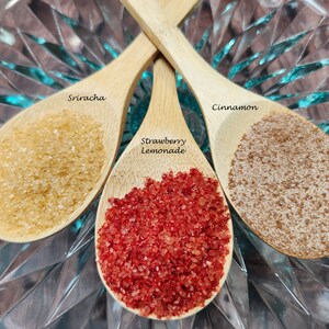 May include: Three wooden spoons filled with different colored sugar crystals. The spoons are labeled "Sriracha", "Strawberry Lemonade", and "Cinnamon".