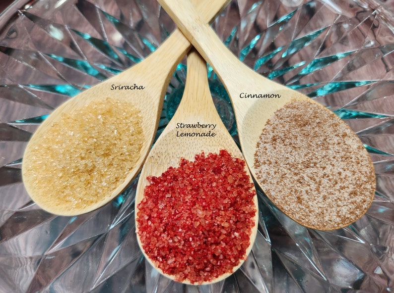 May include: Three wooden spoons filled with different colored sugar crystals. The spoons are labeled "Sriracha", "Strawberry Lemonade", and "Cinnamon".