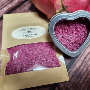 May include: A small heart-shaped tin filled with pink dragon fruit flavored sugar. The tin is sitting on a wooden surface next to a brown paper bag filled with the same sugar. The bag has a label that says "Jazzy T Foods" and "Dragon Fruit Flavored Sugar 1.5 oz."