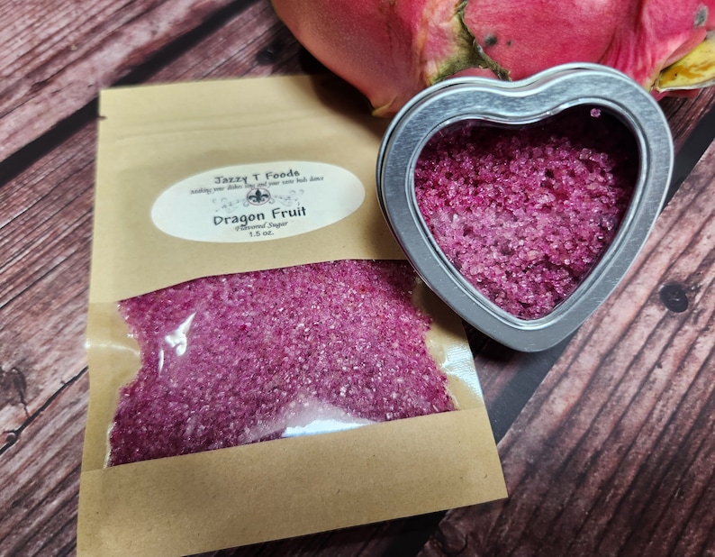 May include: A small heart-shaped tin filled with pink dragon fruit flavored sugar. The tin is sitting on a wooden surface next to a brown paper bag filled with the same sugar. The bag has a label that says "Jazzy T Foods" and "Dragon Fruit Flavored Sugar 1.5 oz."