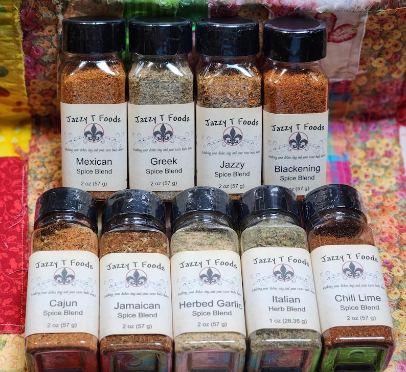 May include: A selection of spice blends in clear glass jars with black lids. The labels read "Jazzy T Foods" and list various spice blends including Mexican, Greek, Cajun, and Italian. Each jar contains 57 g of spice.
