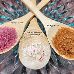 May include: Three wooden spoons filled with different flavored sugar crystals. The spoons are arranged in a triangle shape on a clear glass surface. The flavors are Cherry Almond, White Chocolate Peppermint, and Pumpkin Pie.