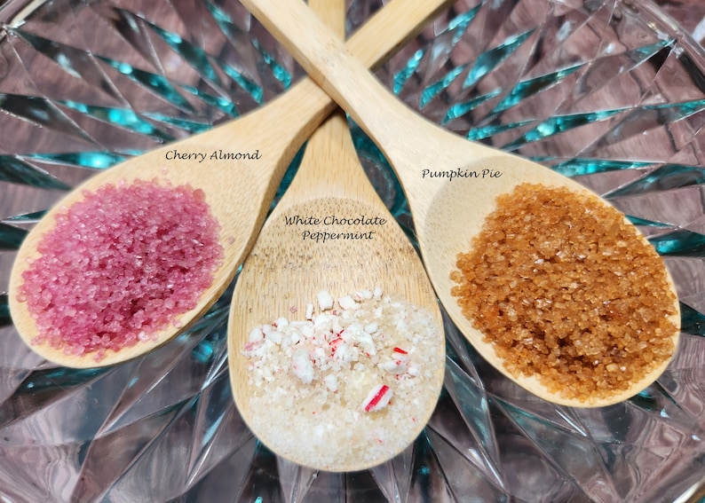 May include: Three wooden spoons filled with different flavored sugar crystals. The spoons are arranged in a triangle shape on a clear glass surface. The flavors are Cherry Almond, White Chocolate Peppermint, and Pumpkin Pie.
