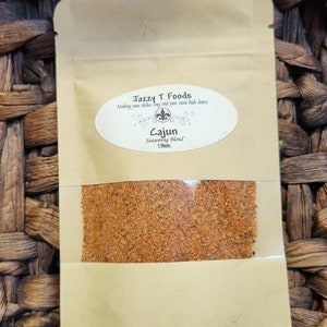 May include: A brown paper bag with a clear window showing a red and orange spice blend. The bag has a white label with the text "Jazzy T Foods" and "Making your dishes sing and your taste buds dance". The label also includes the text "Cajun Seasoning Blend 1.5oz."