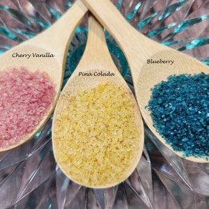 May include: Three wooden spoons filled with colorful sugar crystals. The spoons are labeled "Cherry Vanilla", "Pina Colada", and "Blueberry".