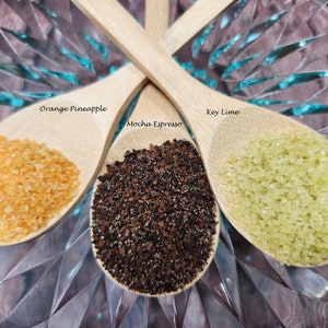 May include: Three wooden spoons filled with different ingredients. The spoon on the left is filled with orange-colored sugar crystals labeled "Orange-Pineapple". The spoon on the right is filled with green sugar crystals labeled "Key Lime". The spoon in the middle is filled with dark brown coffee grounds labeled "Mocha Espresso".
