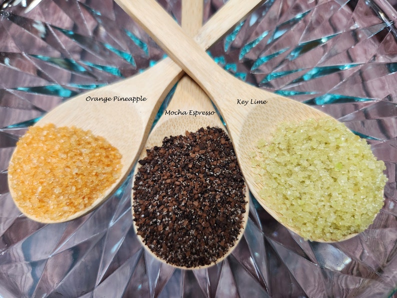 May include: Three wooden spoons filled with different ingredients. The spoon on the left is filled with orange-colored sugar crystals labeled "Orange-Pineapple". The spoon on the right is filled with green sugar crystals labeled "Key Lime". The spoon in the middle is filled with dark brown coffee grounds labeled "Mocha Espresso".