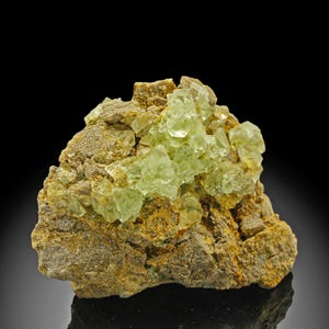 Può includere: Primo piano di un campione minerale con un gruppo di cristalli traslucidi verde pallido su una matrice ruvida di colore marrone chiaro. I cristalli hanno una struttura cubica e la matrice ha una superficie strutturata con sfumature di giallo e marrone.