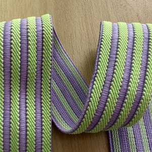 Op de afbeelding: Close-up van een decoratief lint met een herhalend patroon van verticale strepen. De strepen wisselen af tussen een lichtgroene en een lavendelkleur. Het lint is gemaakt van een geweven materiaal, waarschijnlijk voor knutselen of verfraaien.
