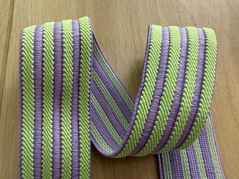 Op de afbeelding: Close-up van een decoratief lint met een herhalend patroon van verticale strepen. De strepen wisselen af tussen een lichtgroene en een lavendelkleur. Het lint is gemaakt van een geweven materiaal, waarschijnlijk voor knutselen of verfraaien.
