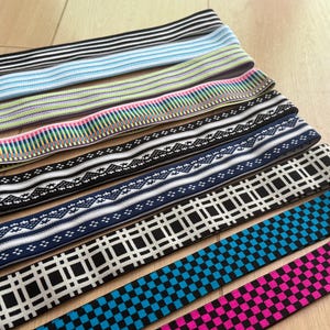 Op de afbeelding: Assortiment elastische banden in diverse kleuren en patronen. Ontwerpen omvatten strepen, geometrische vormen en bloemmotieven. Kleuren vari&euml;ren van zwart en wit tot blauw, roze en groen. De banden zijn waarschijnlijk voor naai- of knutselprojecten.