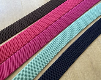 May include: Four different colors of elastic webbing, including black, brown, mint green, and pink. The webbing has a textured surface and is shown lying flat on a wooden surface.