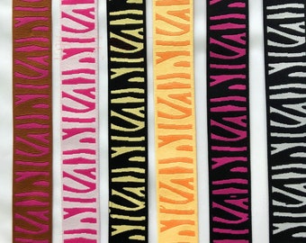 Puede incluir: Seis colores diferentes de ribete elástico con un patrón repetido de una letra estilizada "M". Los colores son marrón, rosa, amarillo, naranja, magenta y gris.