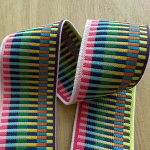 Op de afbeelding: Een kleurrijke elastische band met een geometrisch patroon. De band heeft horizontale strepen in roze, paars, blauw, groen, geel en oranje. De randen zijn wit en de band is over zichzelf gevouwen.