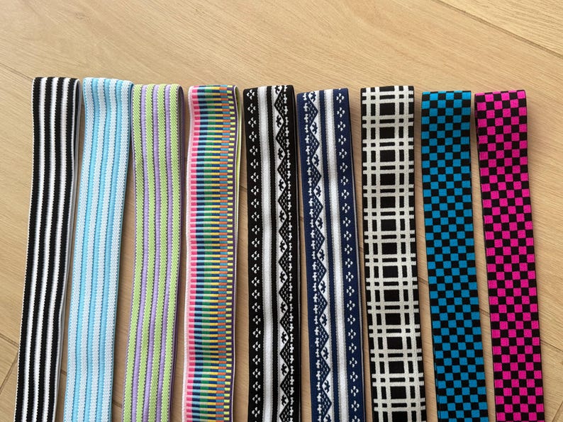 Op de afbeelding: Een verzameling elastische banden in verschillende patronen en kleuren. Ontwerpen omvatten zwart-witte strepen, blauwe strepen, groen-paarse strepen, een regenboogpatroon en zwart-witte geometrische vormen.