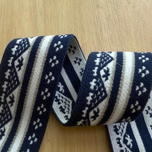 Op de afbeelding: Een lengte geweven lint met een herhalend patroon. Het lint heeft een marineblauwe achtergrond met witte en lichtbruine geometrische ontwerpen. Het lint is opgerold en wordt gepresenteerd op een licht houten oppervlak.