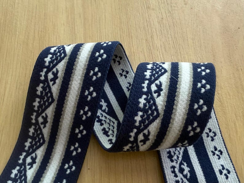 Op de afbeelding: Een lengte geweven lint met een herhalend patroon. Het lint heeft een marineblauwe achtergrond met witte en lichtbruine geometrische ontwerpen. Het lint is opgerold en wordt gepresenteerd op een licht houten oppervlak.