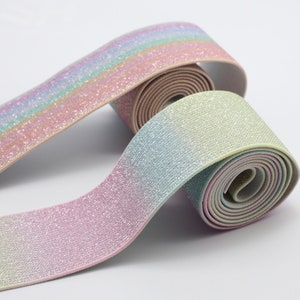 Op de afbeelding: Twee rollen elastisch lint in een verloop van pastelkleuren. Het lint is glinsterend en lijkt gemaakt van een zacht, flexibel materiaal. De kleuren gaan over van roze naar blauw en groen.