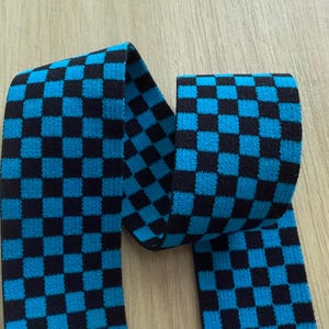 Op de afbeelding: Een stuk stof met een zwart en turkooisblauw ruitpatroon. De stof is gevouwen en ligt op een licht houten oppervlak. Het patroon bestaat uit gelijkmatig verdeelde vierkanten.