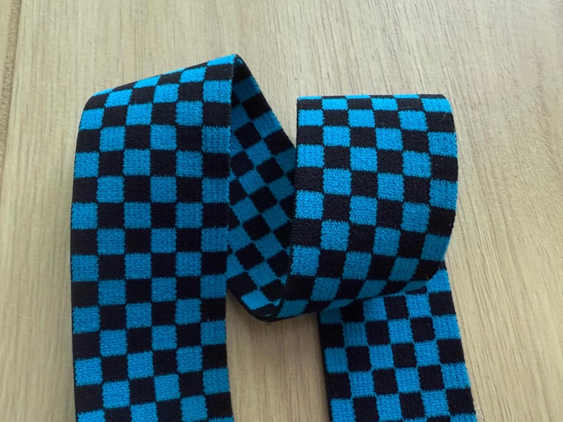 Op de afbeelding: Een stuk stof met een zwart en turkooisblauw ruitpatroon. De stof is gevouwen en ligt op een licht houten oppervlak. Het patroon bestaat uit gelijkmatig verdeelde vierkanten.