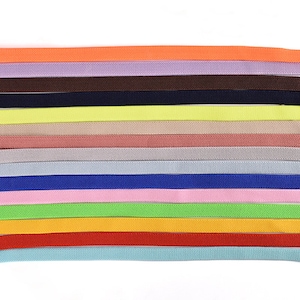 Pode incluir: Uma coleção de 15 elásticos de cores diferentes, cada um com aproximadamente 1,25 cm de largura. As cores incluem laranja, lavanda, marrom, preto, amarelo, pêssego, cinza, azul claro, rosa, verde limão, amarelo, vermelho e azul claro.