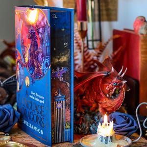 Op de afbeelding: Een blauw hardback boek getiteld "A Language of Dragons" door S.A. Williamson. De boekomslag toont een draak met rode, oranje en gele vlammen. Het boek ligt op een houten tafel met een brandende kaars en een drakenbeeldje.