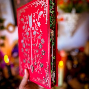 May include: A red book with silver floral and arrow designs on the cover. The spine features green and red ivy-like patterns. The book's edges are gilded, and the overall design suggests a fantasy or historical theme.