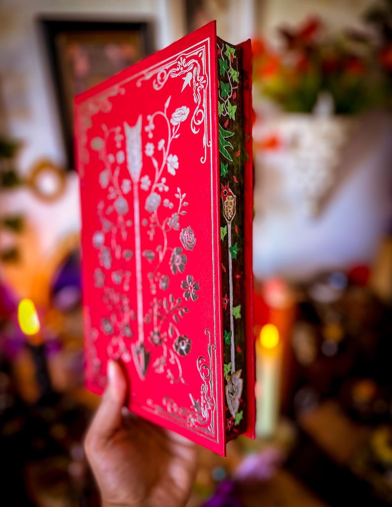 May include: A red book with silver floral and arrow designs on the cover. The spine features green and red ivy-like patterns. The book's edges are gilded, and the overall design suggests a fantasy or historical theme.